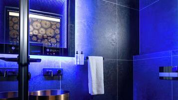 Un baño moderno con azulejos azul oscuro, un espejo grande, un lavabo redondo y un inodoro.
