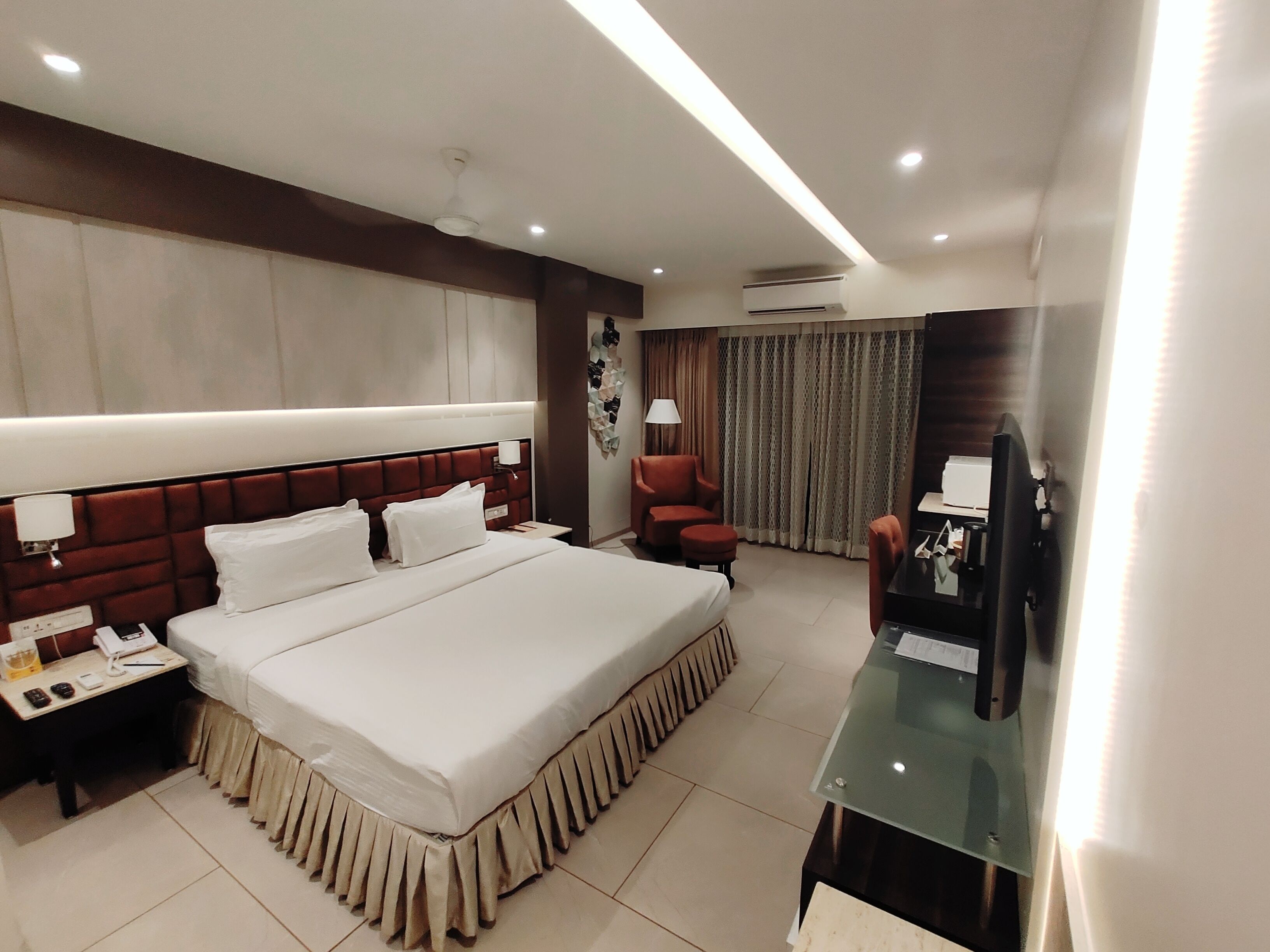 A hotel room with a large bed, a desk with a computer, a chair, and an air conditioning unit.