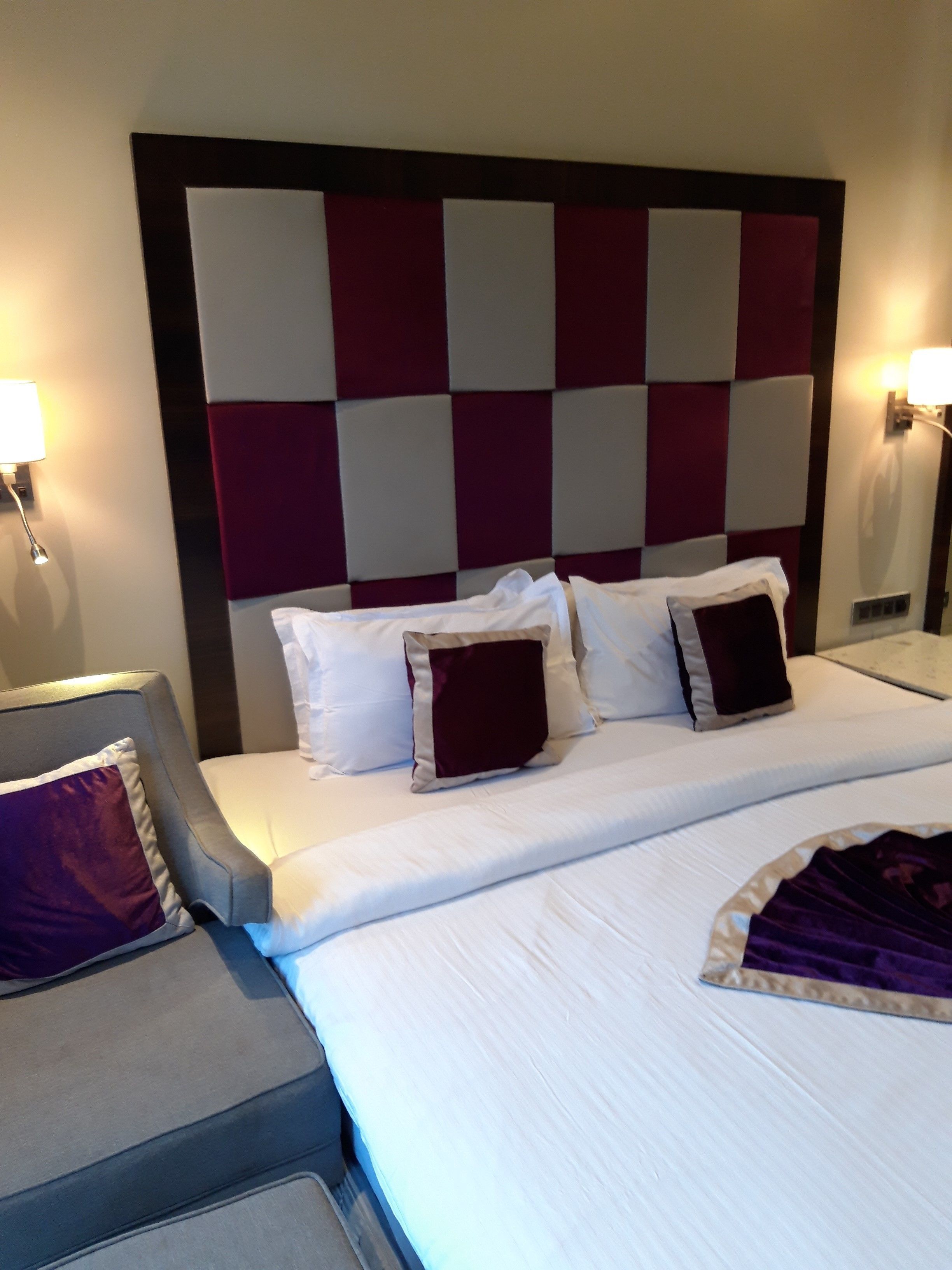 Quarto de hotel com uma cama grande, uma poltrona cinzenta e uma mesa de cabeceira com abajur.