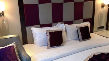 Quarto de hotel com uma cama grande, uma poltrona cinzenta e uma mesa de cabeceira com abajur.