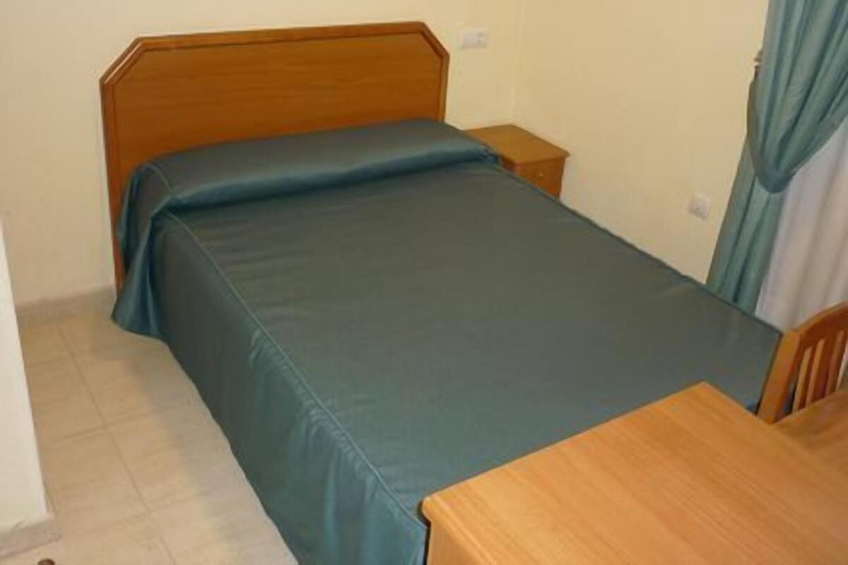 Ein kleines Zimmer mit einem Einzelbett, einem Holzkopfstück, einem Nachttisch aus Holz und einem Holztisch.