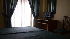 A hotel room with a bed, a television on a wooden stand, a chair, and a window with curtains. - Hotel Tuto (Torrevieja)