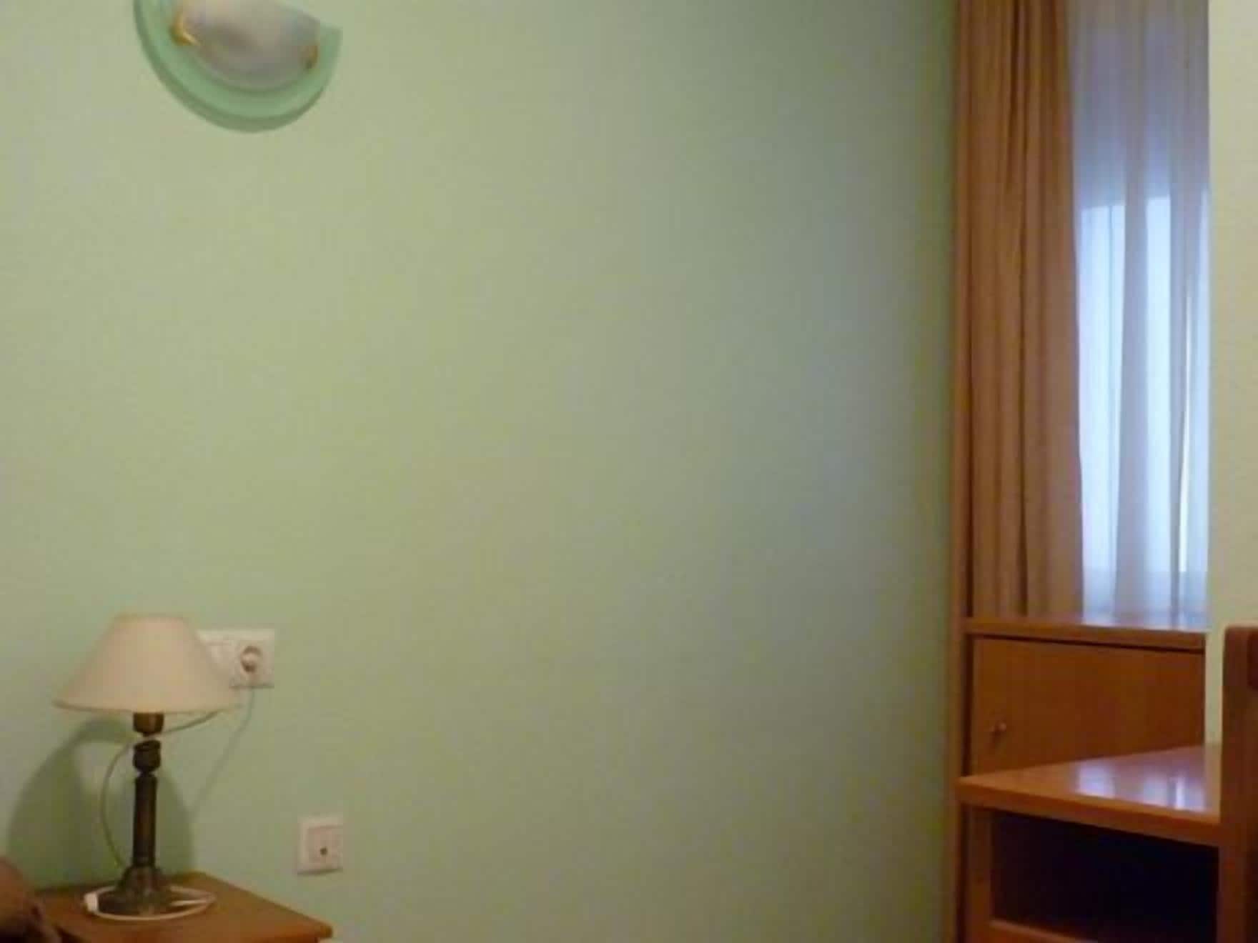Une pièce avec un mur vert, une table de chevet en bois avec une lampe, une fenêtre avec des rideaux et une étagère en bois.
