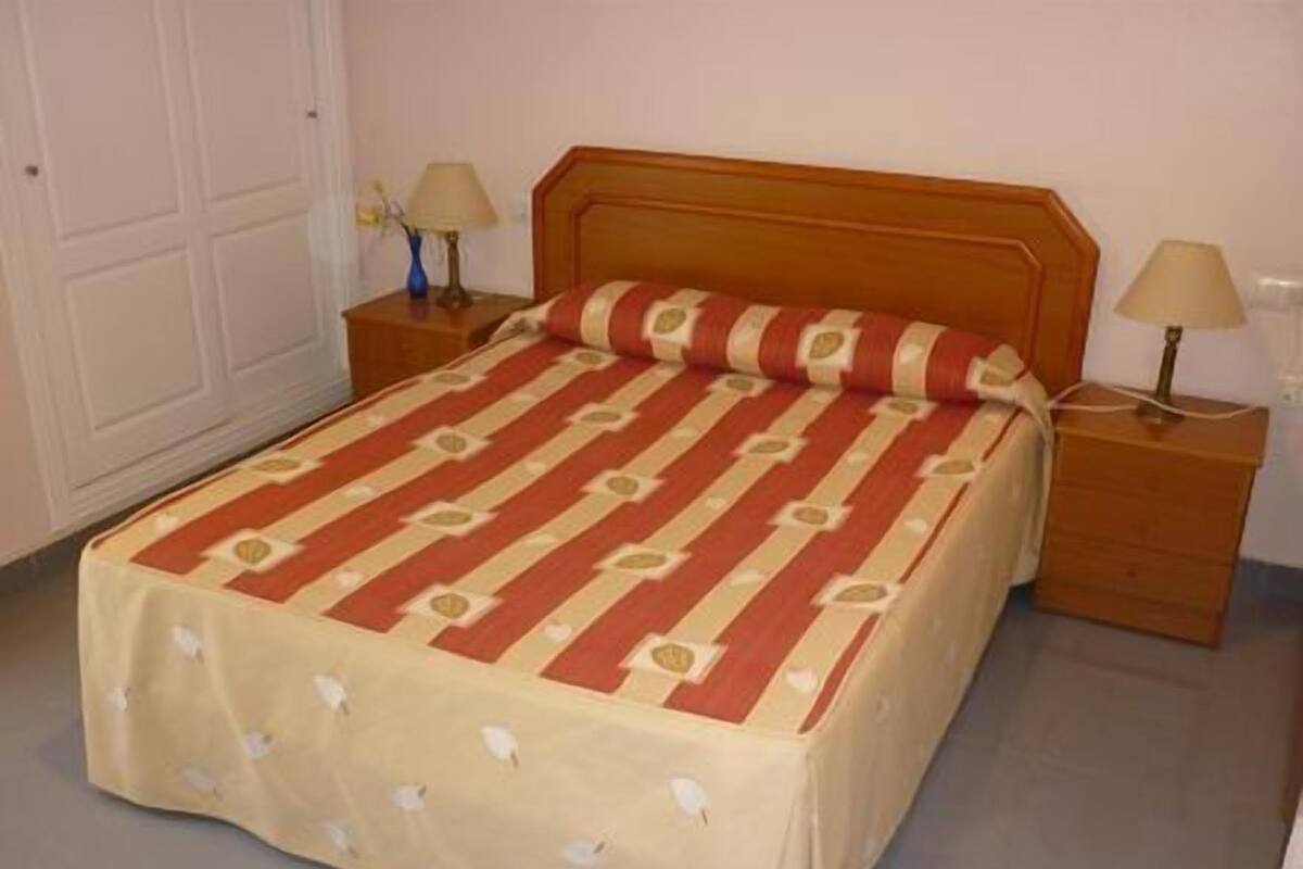 Ein ordentlich bezogenes Bett mit gestreifter Bettdecke, zwei Nachttische mit Lampen und eine schlichte Wand.