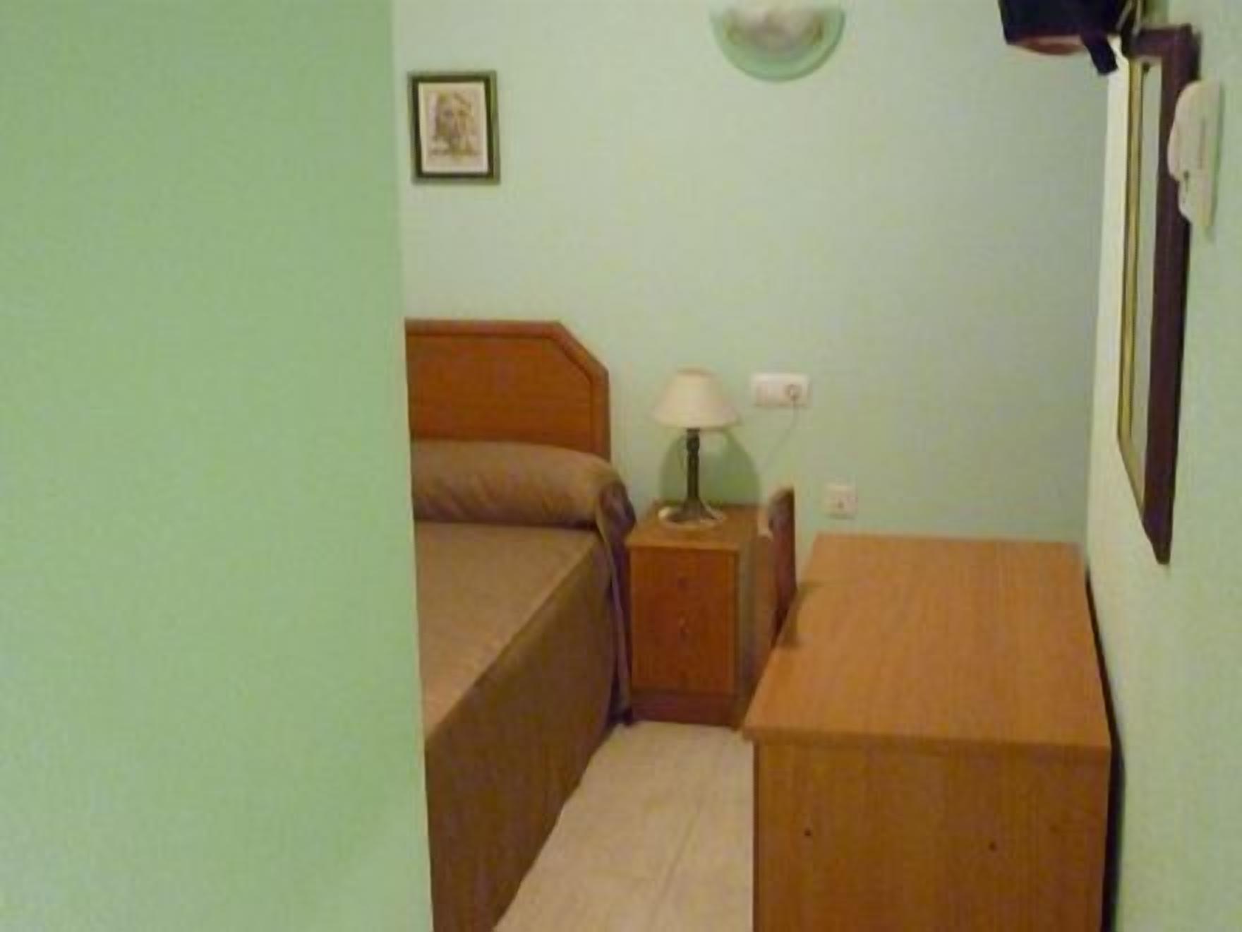 A small, simple room with a bed, a nightstand with a lamp, and a wooden dresser.