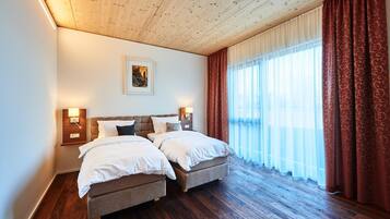 Une chambre d’hôtel avec deux lits, un plancher en bois, un plafond avec des poutres apparentes, une grande fenêtre avec des rideaux et des lampes fixées au mur.