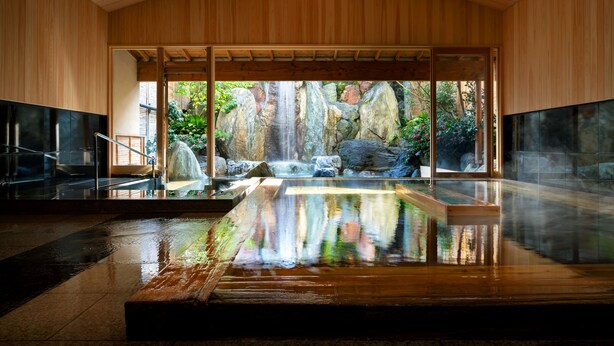 Un onsen japonés tradicional con interior de madera, una fuente termal de piedra y vistas a una formación rocosa natural.