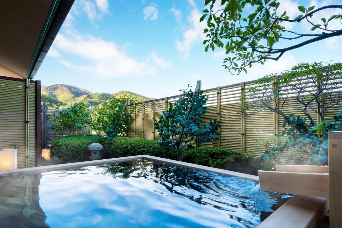 A hot spring bath area with a wooden deck, bamboo fence, and lush greenery.