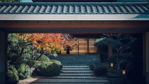 Edificio japonés tradicional con tejado de tejas, un sendero de piedra que conduce a una puerta de madera y un jardín con follaje otoñal.