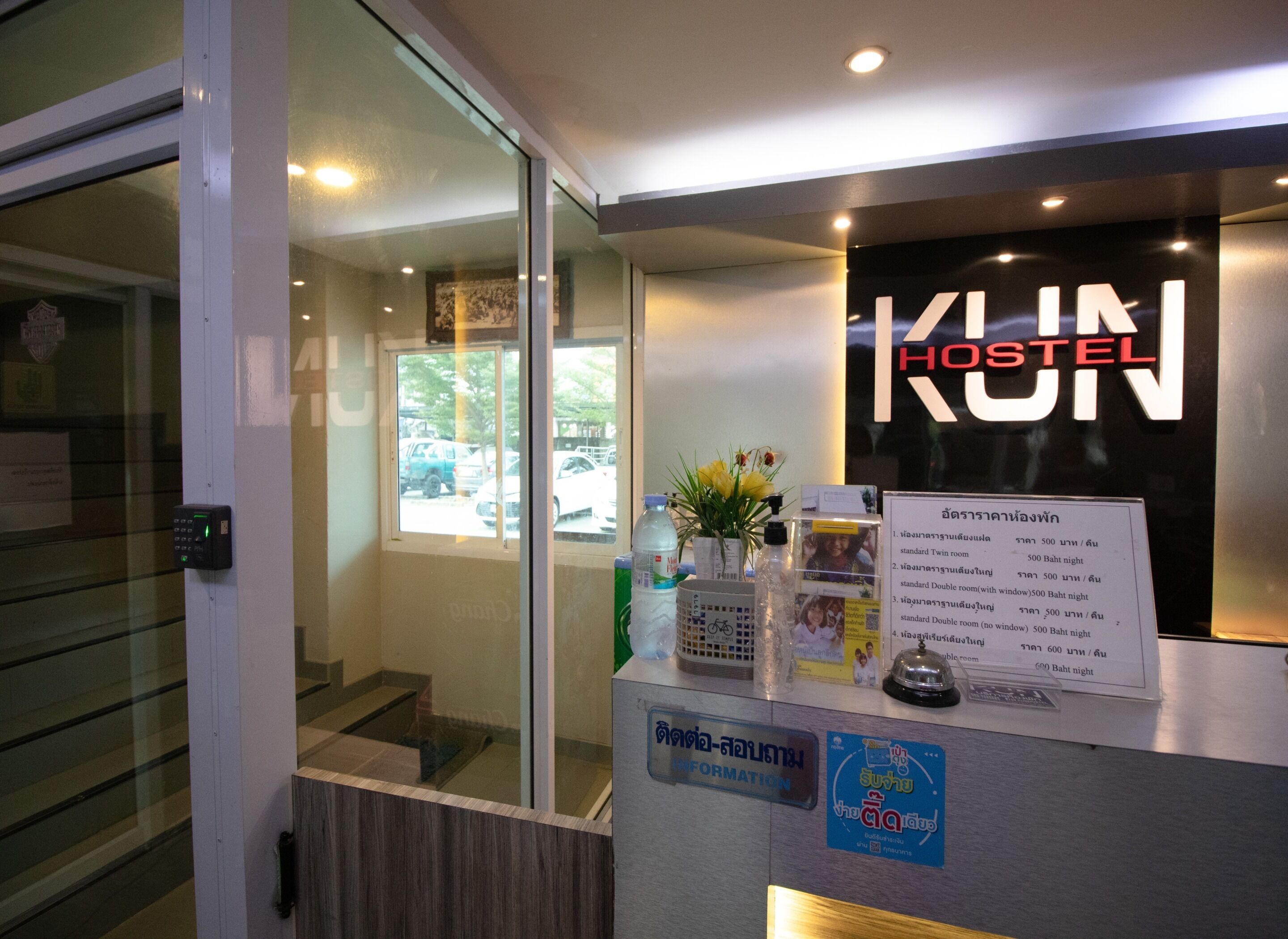 A reception desk with a sign reading "KUN HOSTEL" and a list of room rates in Thai.
