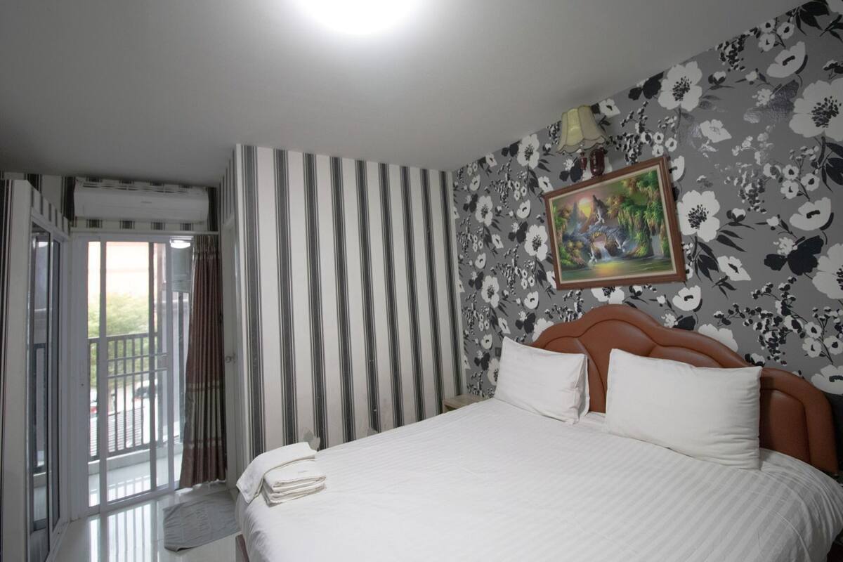 Una habitación de hotel con cama, ventana con cortinas, pared con estampado floral y unidad de aire acondicionado.