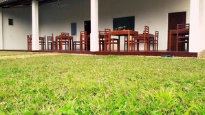 A covered outdoor area with a lawn, a wooden table with chairs, and a building with white walls and windows. - Sinharaja Forest Sanctuary Hotel (Deniyaya)
