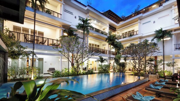 Un hotel de varios pisos con piscina, zona de estar exterior y abundante vegetación.