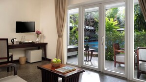 Habitación de hotel con televisión de pantalla plana, un escritorio con un florero, una cesta de frutas y vistas a una piscina y jardín al aire libre.