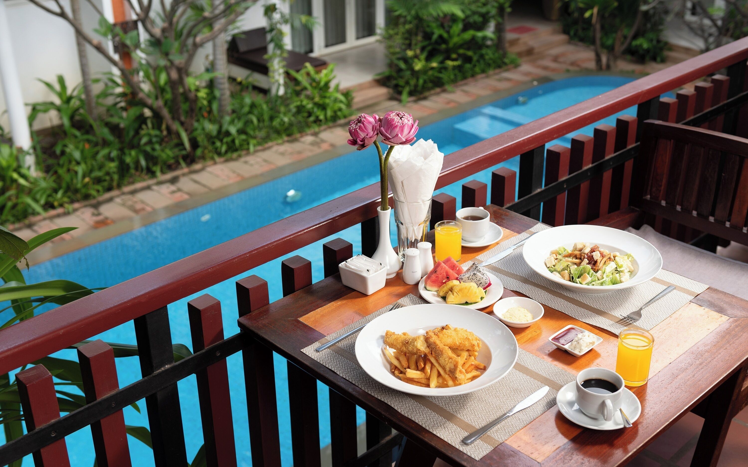 A dining table with a meal, a vase with flowers, and a view of a swimming pool.