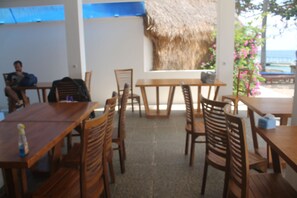 An indoor dining area with wooden tables and chairs, a thatched wall, and a view of the sea. - The Place Beach Bungalows (Sekotong Barat)