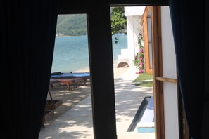 A view through a window of a pool area with a bench and a table, and a building with a white exterior and a wooden deck. - The Place Beach Bungalows (Sekotong Barat)