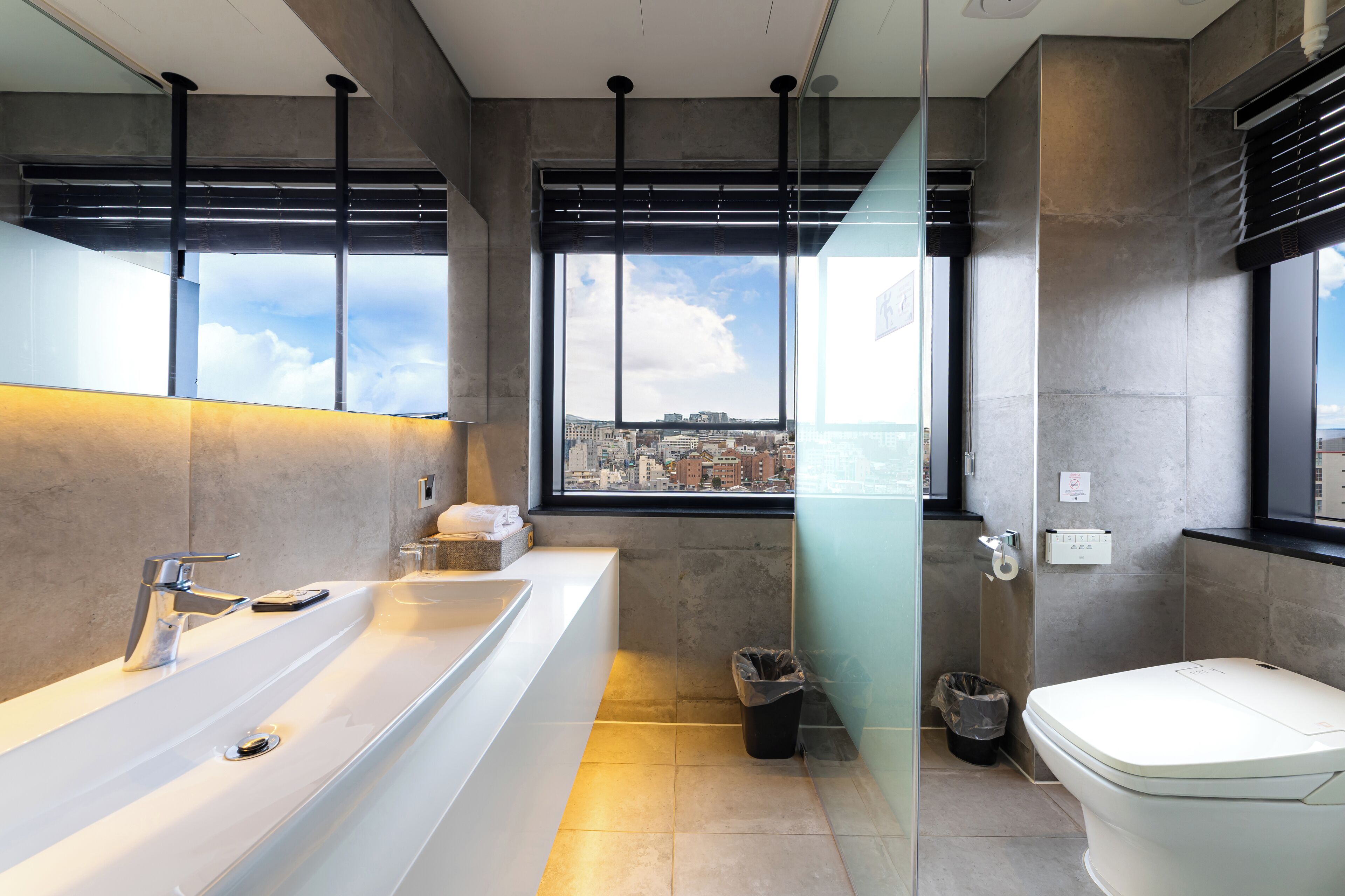 A modern bathroom with a large bathtub, a toilet, and a window offering a city view.