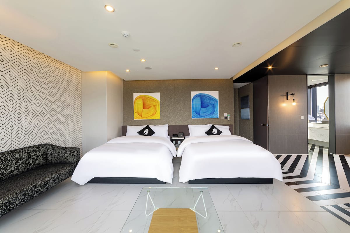 Habitació d'hotel moderna amb dos llits, un sofà, una taula de cafè de vidre i una obra d'art abstracta a la paret.