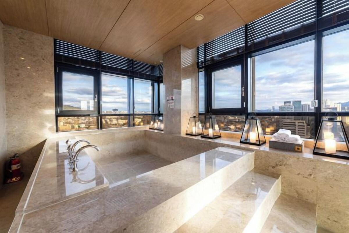 A modern bathroom with a large bathtub, a sink, and a window with a city view.