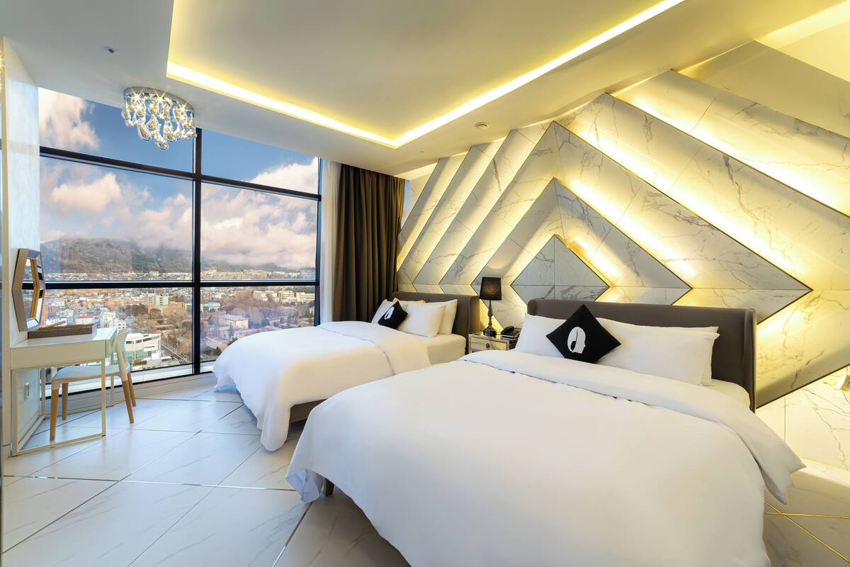 Habitació d'hotel moderna amb dos llits, un escriptori i una finestra gran que ofereix vistes a la ciutat.