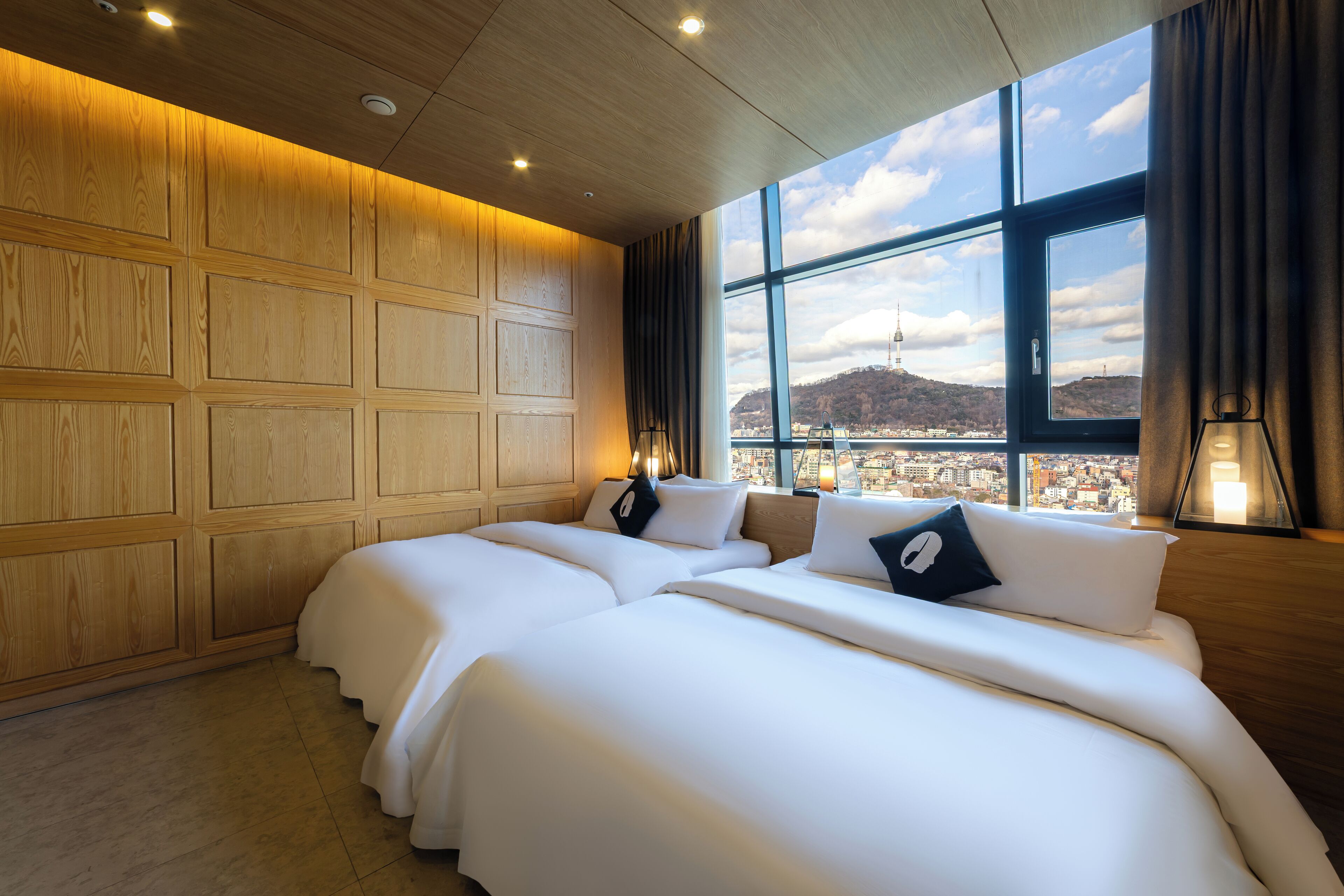 A hotel room with three beds, wooden paneling on the walls, and a large window offering a city view.