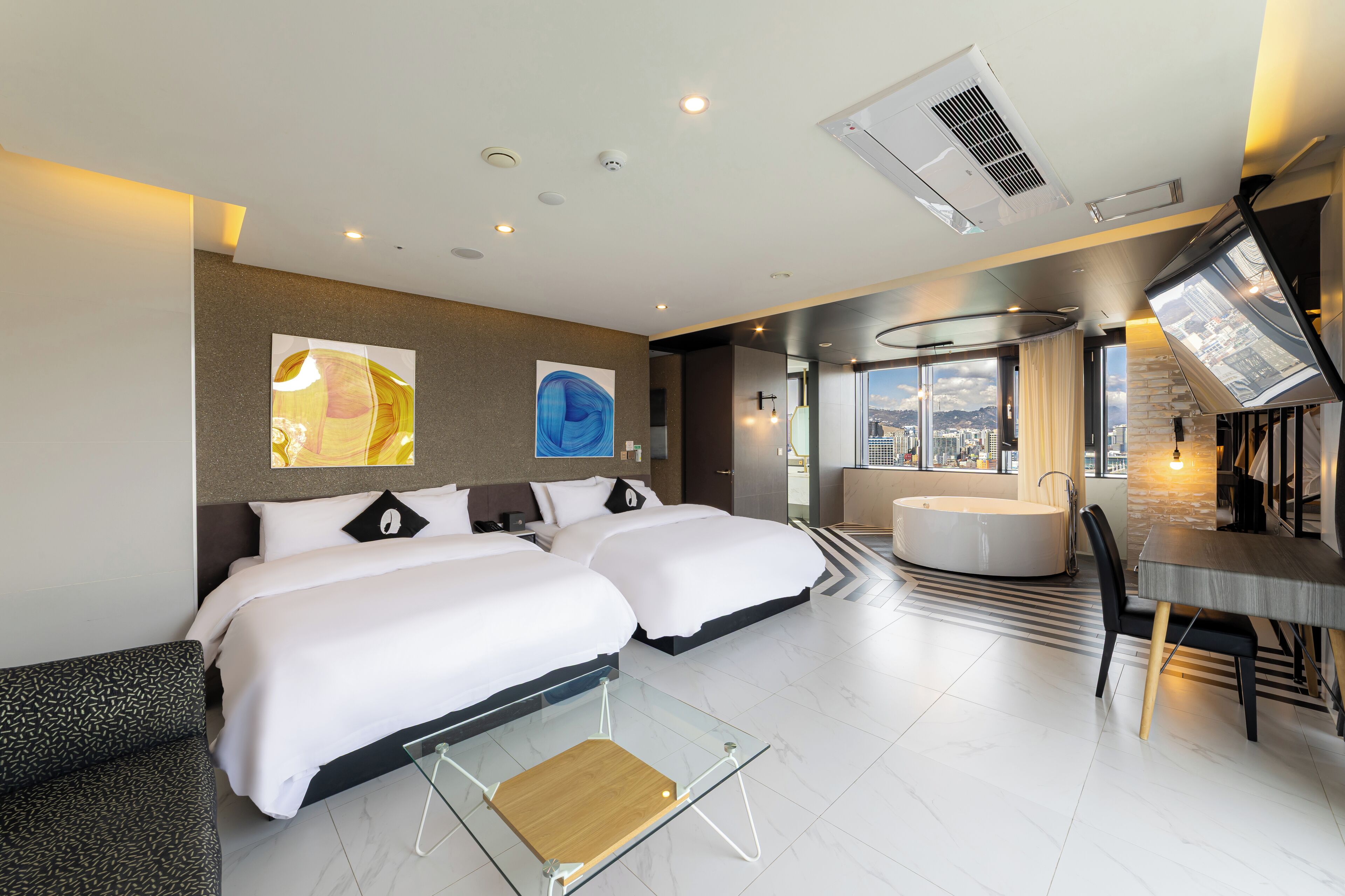 A modern hotel room with two large beds, a glass coffee table, a dining area, and a view of the city.