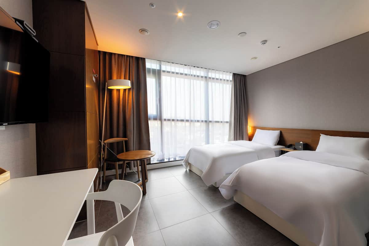 Habitació d'hotel amb dos llits, un escriptori, una cadira, una televisió i una finestra amb cortines.