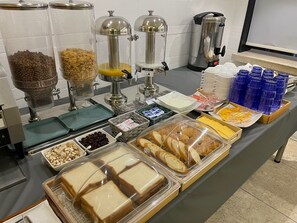 Een ontbijtbuffet met diverse gerechten en dranken, zoals ontbijtgranen, brood en een koffieapparaat.
