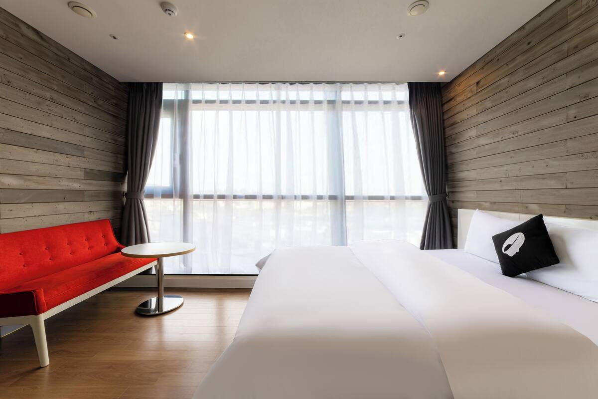 Habitació d'hotel amb un sofà vermell, un llit blanc i una taula rodona petita.