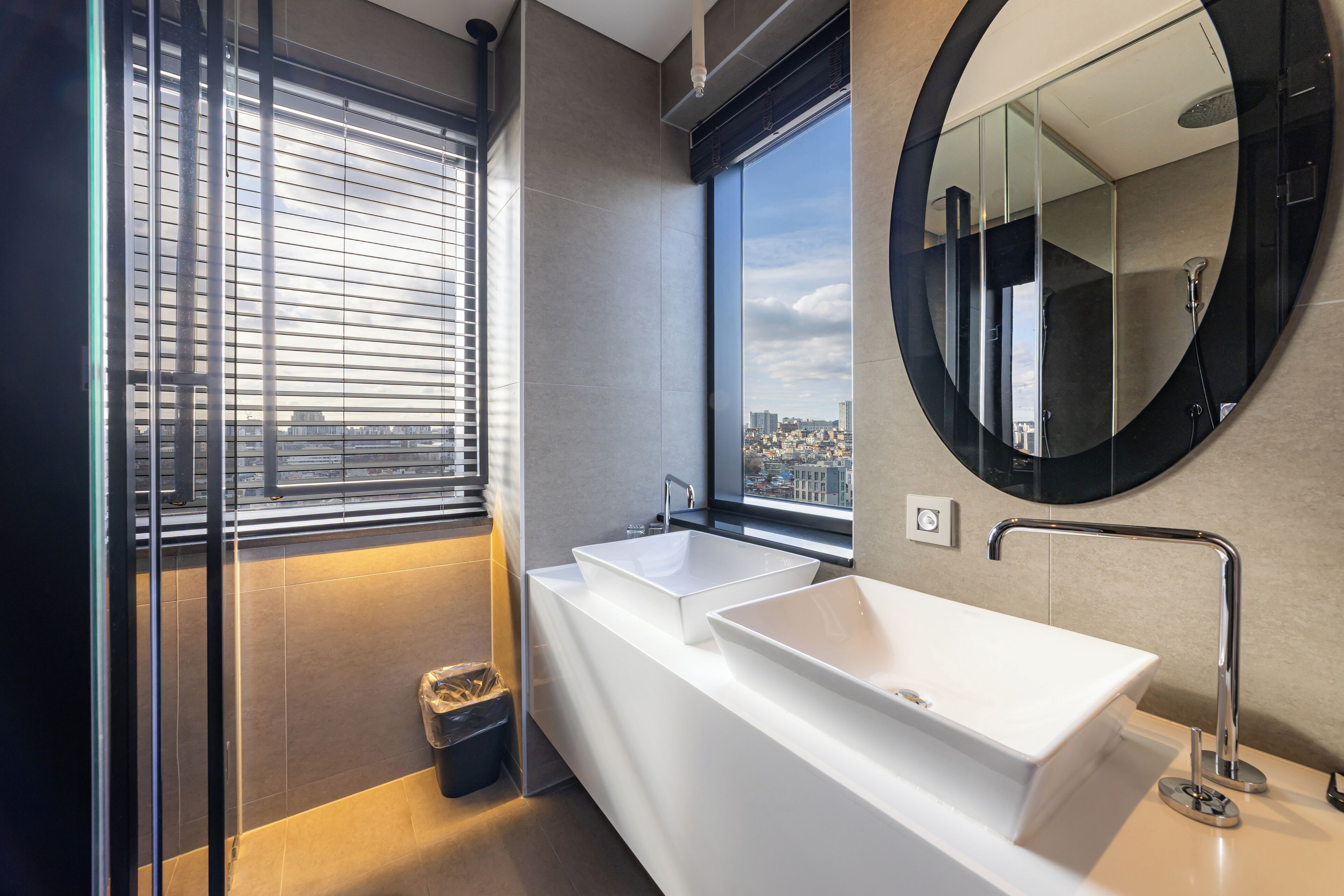 A modern bathroom with a large window, two sinks, and a round mirror.