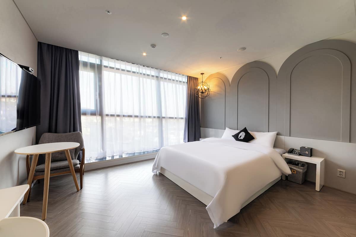 Habitació d'hotel moderna amb un llit gran, una petita taula de treball, una cadira i una finestra gran amb cortines.