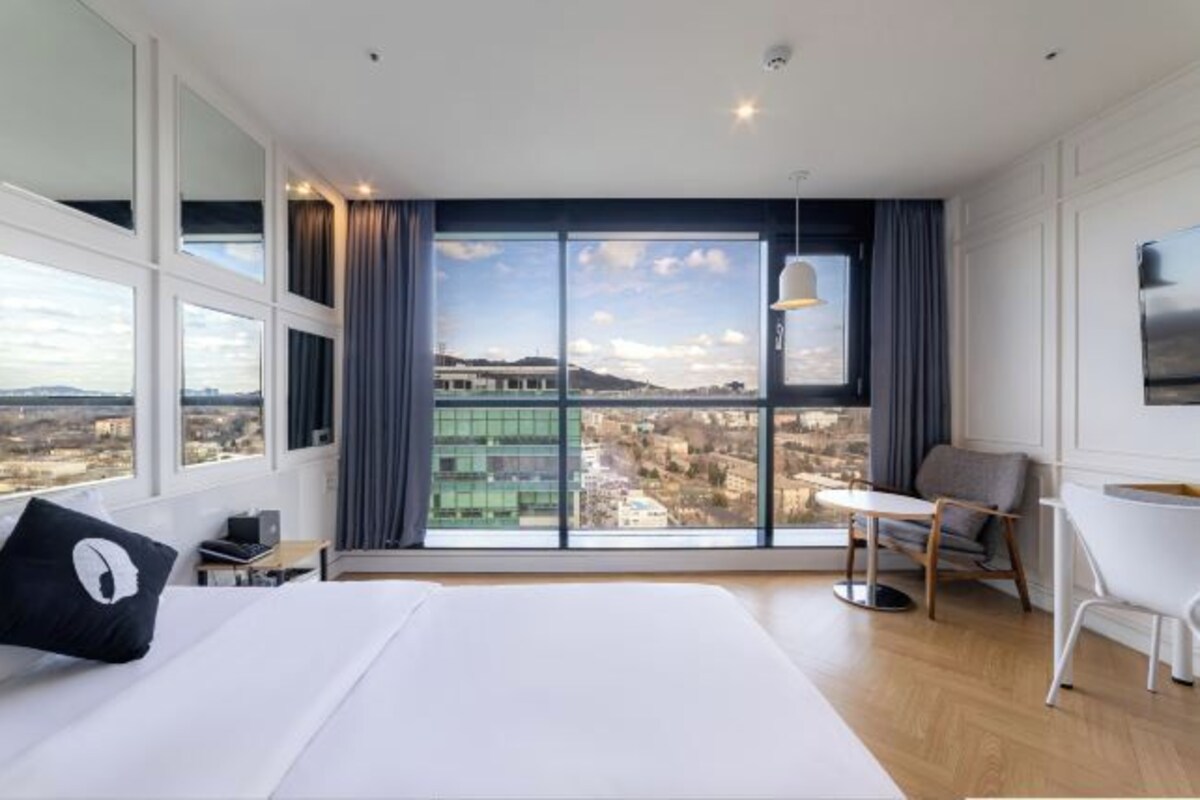 A modern hotel room with a large window offering a city view, a bed with white bedding, a small table, and a chair.