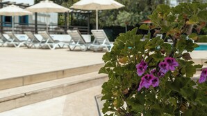 Zona de piscina con sillones de playa y sombrillas, un edificio al fondo y una planta con flores moradas en primer plano.