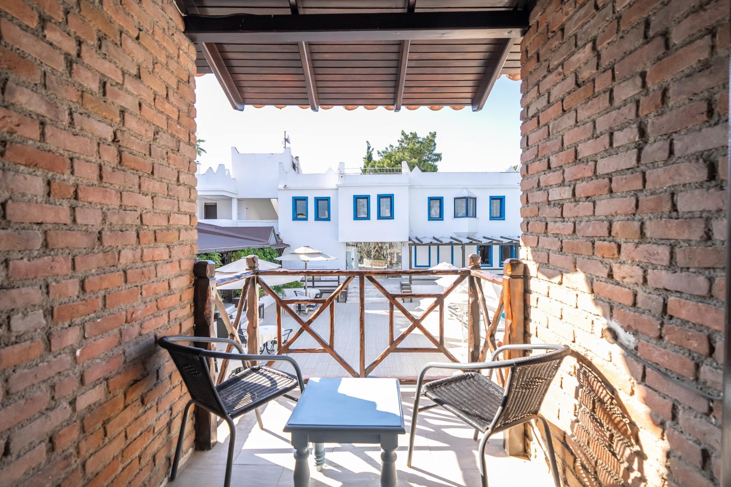 A covered outdoor seating area with brick walls, a wooden railing, and a view of white buildings with blue trim.