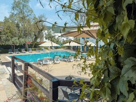 Piscina com espreguiçadeiras e guarda-sóis, rodeada por vegetação e edifícios.