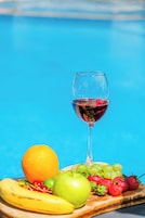 Um copo de vinho tinto, uma laranja, uma banana e várias frutas sobre uma tábua de madeira, com fundo azul.
