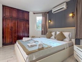 Quarto com cama, guarda-roupa, ar condicionado, mesa de cabeceira e janela com cortinas.