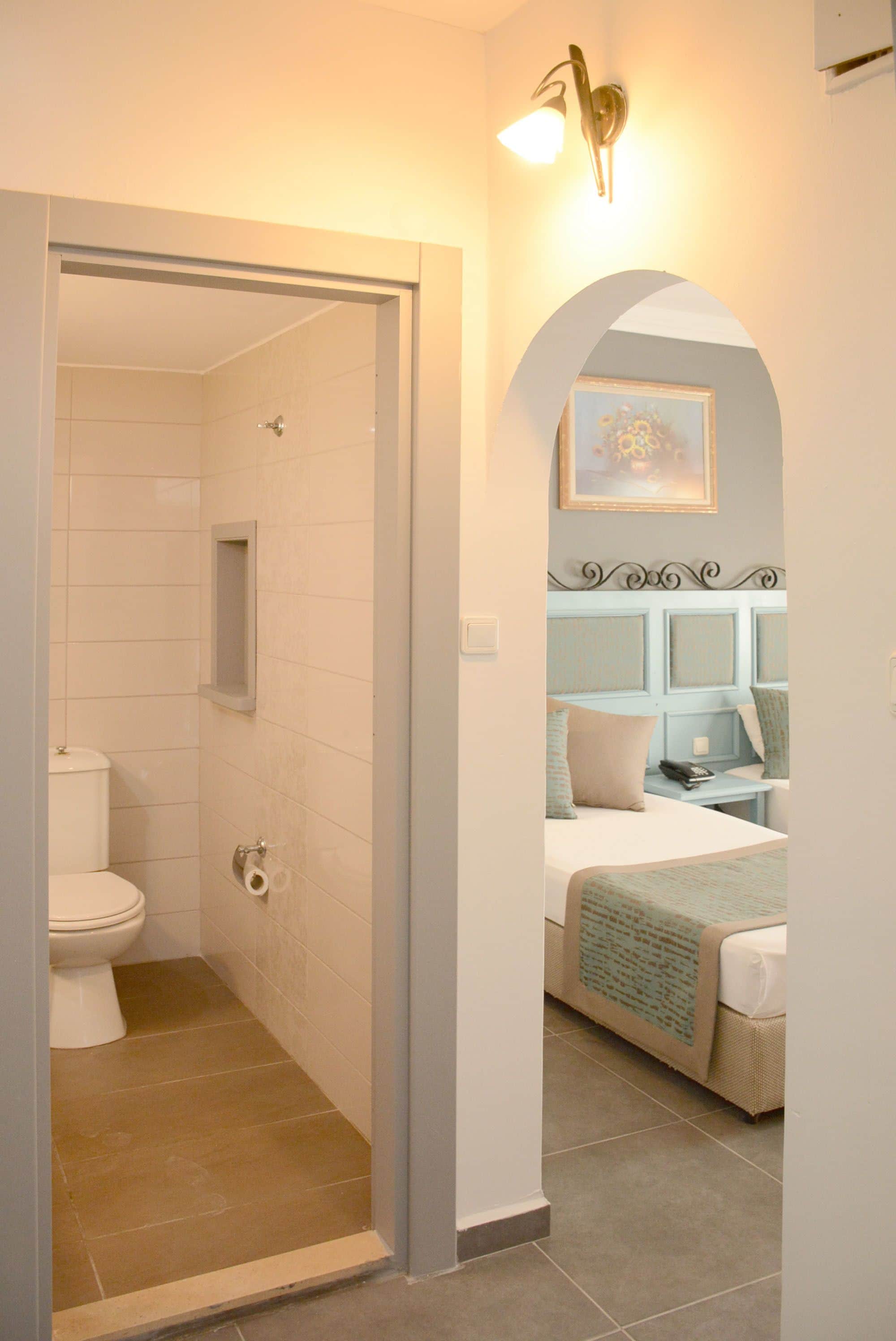 Une salle de bain avec des toilettes, une douche et une petite étagère. Il y a une chambre avec un lit, une table de chevet et un tableau au mur.