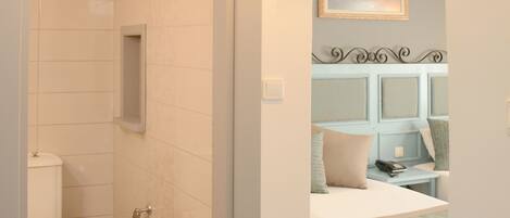 Une salle de bain avec des toilettes, une douche et une petite étagère. Il y a une chambre avec un lit, une table de chevet et un tableau au mur.