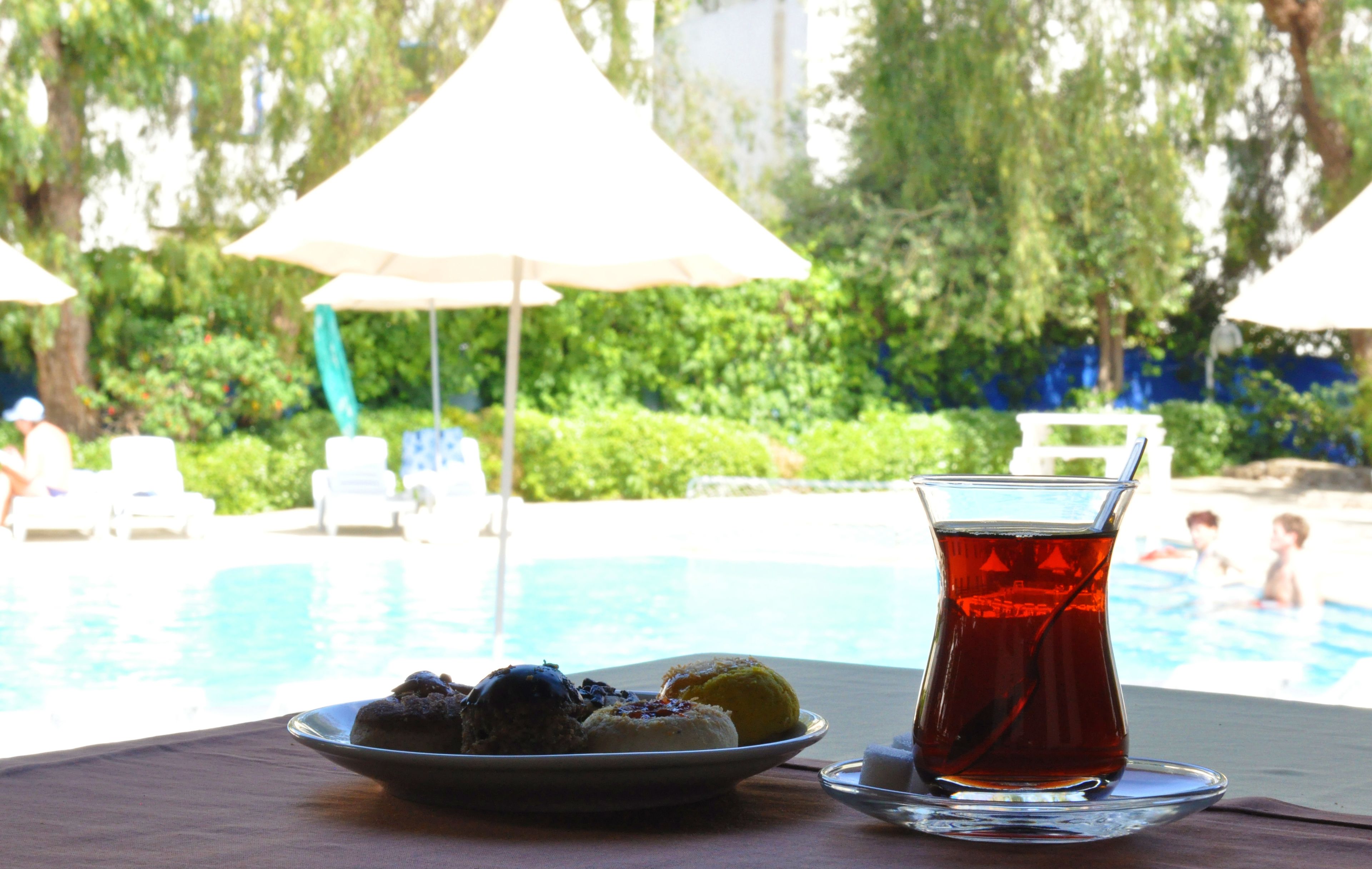 Une table avec une assiette de pâtisseries et une tasse de thé, donnant sur une piscine bordée de parasols et de verdure.