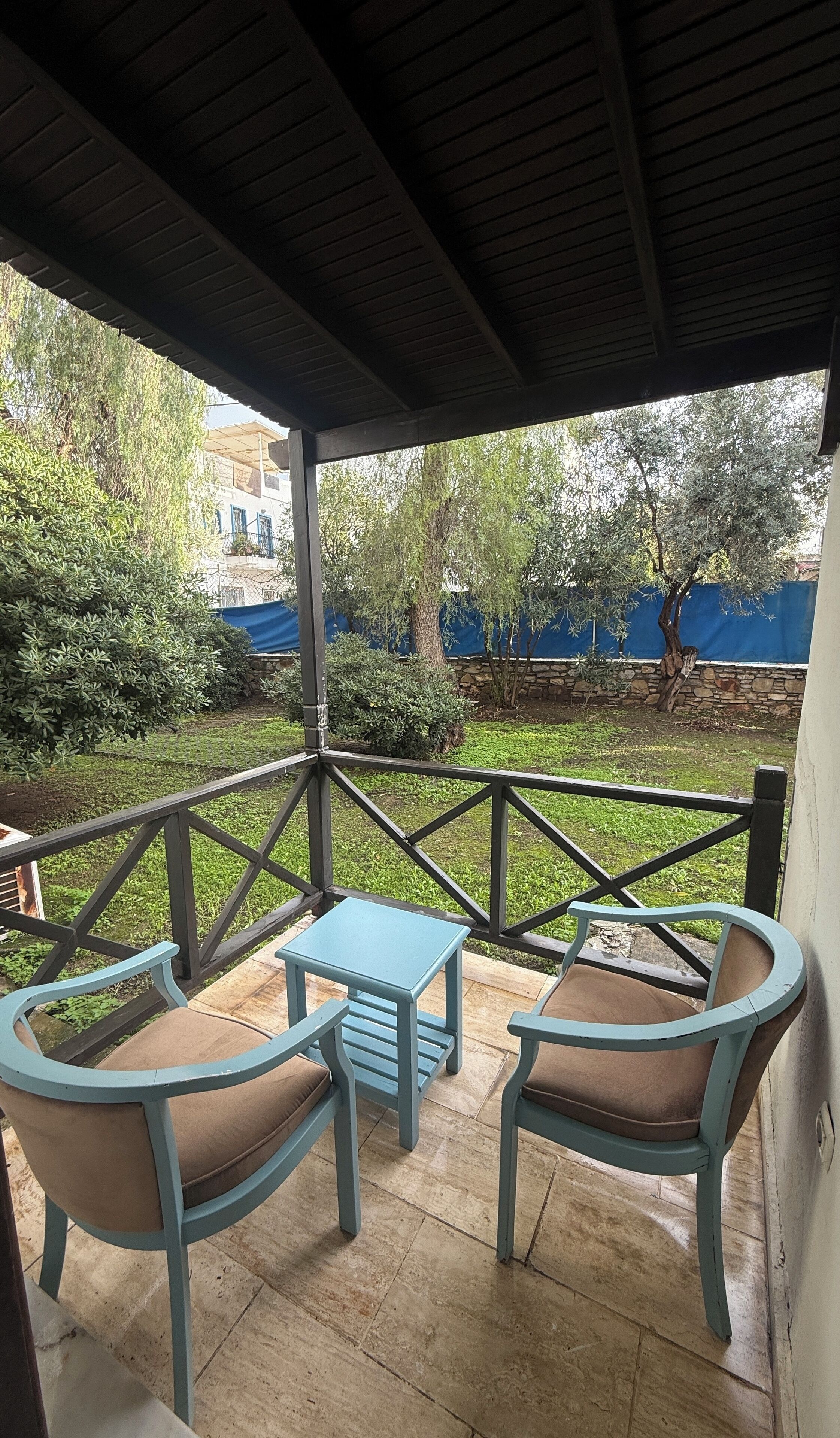 A covered patio area with two chairs and a small table, surrounded by a stone wall and greenery.