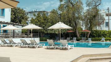 Uma área de piscina com espreguiçadeiras e guarda-sóis, rodeada por árvores e edifícios.