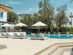 Un área de piscina con sillones de descanso y sombrillas, rodeada de árboles y edificios.