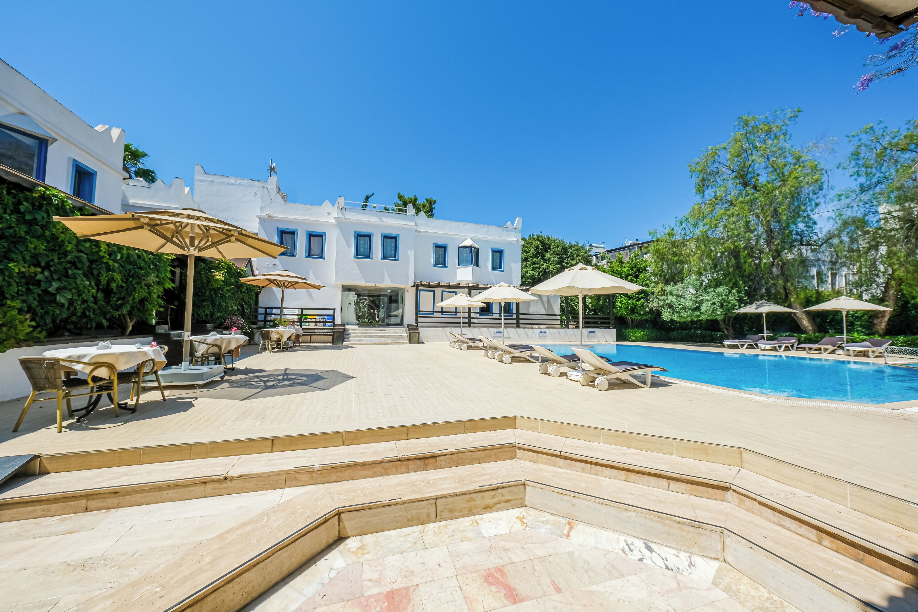 Un espace aménagé au bord de la piscine, avec des chaises longues et des parasols, un ciel d’un bleu limpide et un bâtiment aux murs blancs et aux bordures bleues.