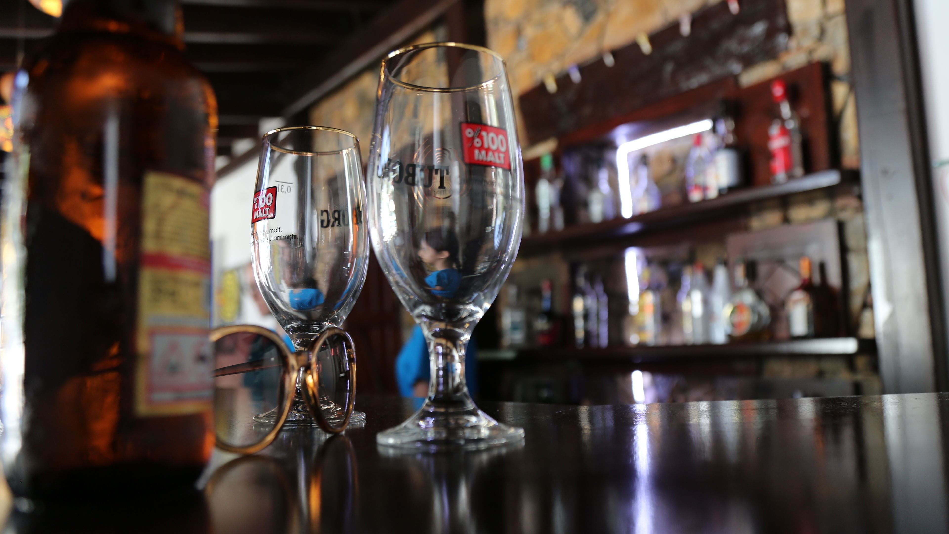 Two wine glasses on a bar counter with a bottle of beer and a shelf of various bottles in the background.
