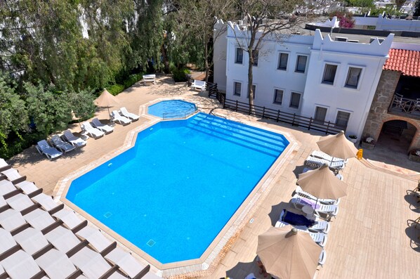 Une grande piscine rectangulaire, bordée de chaises longues et d’ombrelles, entourée d’arbres et d’un bâtiment blanc doté de nombreuses fenêtres.