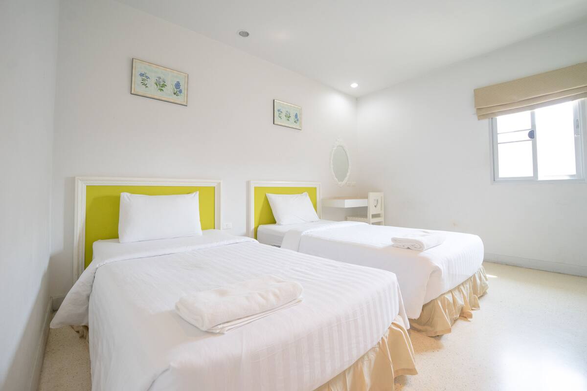 A hotel room with two beds, a window with a beige blind, and two wall-mounted paintings.