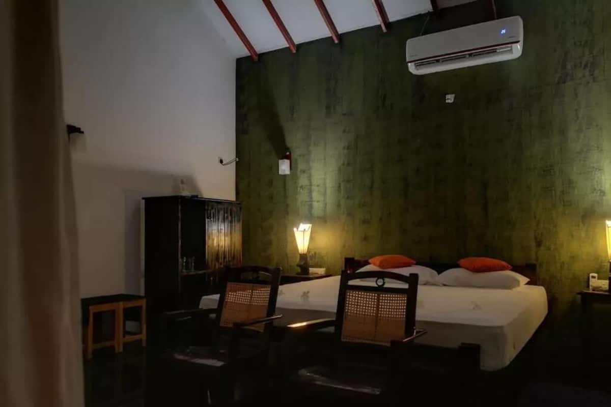 Una habitación con cama, dos sillas, una mesita de noche, una lámpara y aire acondicionado.