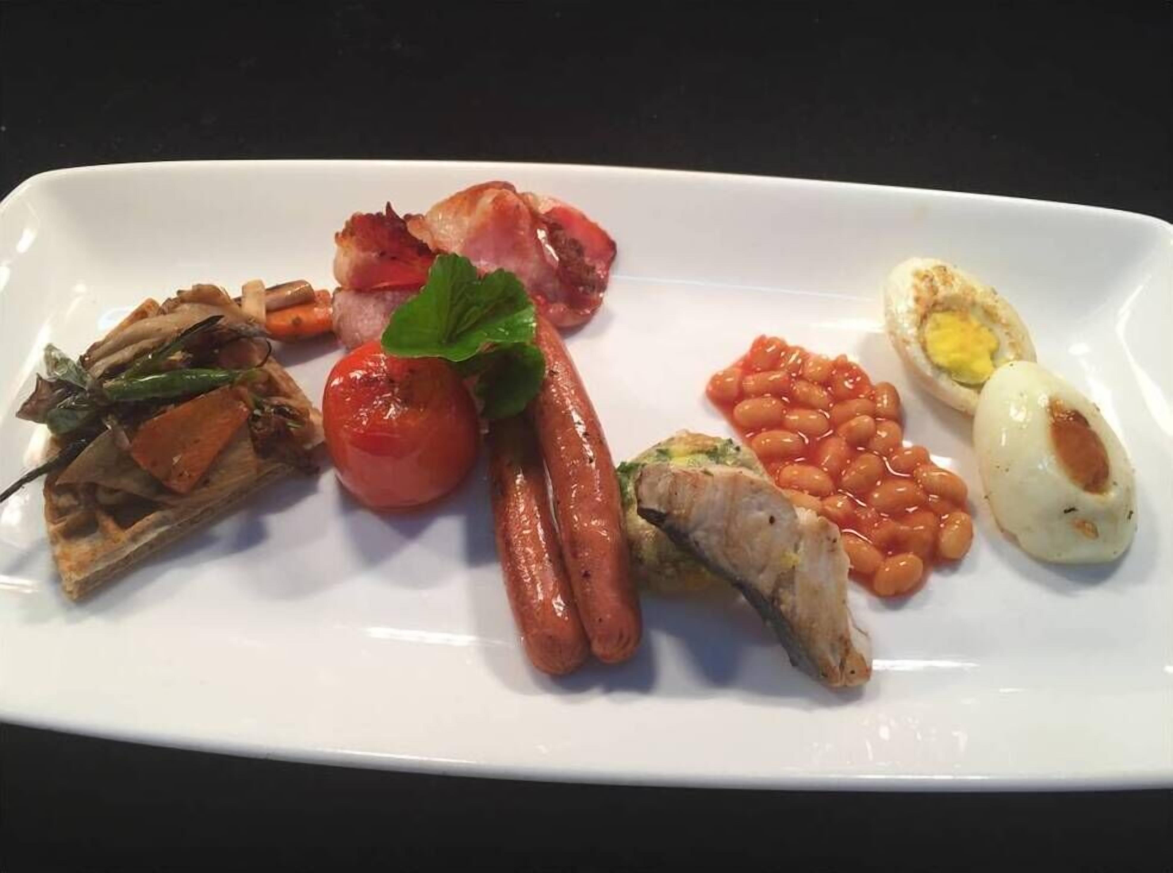 A white rectangular plate with various breakfast items including sausages, eggs, beans, and a tomato.